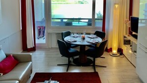 A dining area with a table set for four, a sofa, a TV, and a view of a pool through large windows. - App. 511 by Interhome (Bissone)