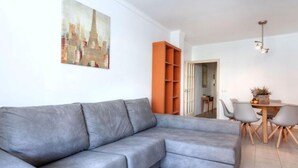 A modern living room with a grey sectional sofa, a wooden dining table with chairs, and a cityscape painting on the wall. - Apartment  Ronda by Interhome (L'Escala)