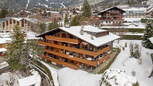 Uma pousada de madeira com vários andares e varandas, situada em uma área montanhosa coberta de neve, com outras construções ao fundo.