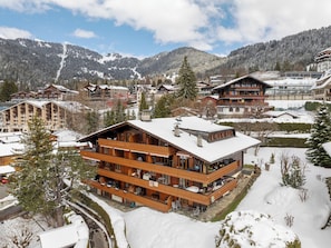 Una cabaña de madera de varios pisos con balcones, ubicada en una zona montañosa nevada con otros edificios al fondo.