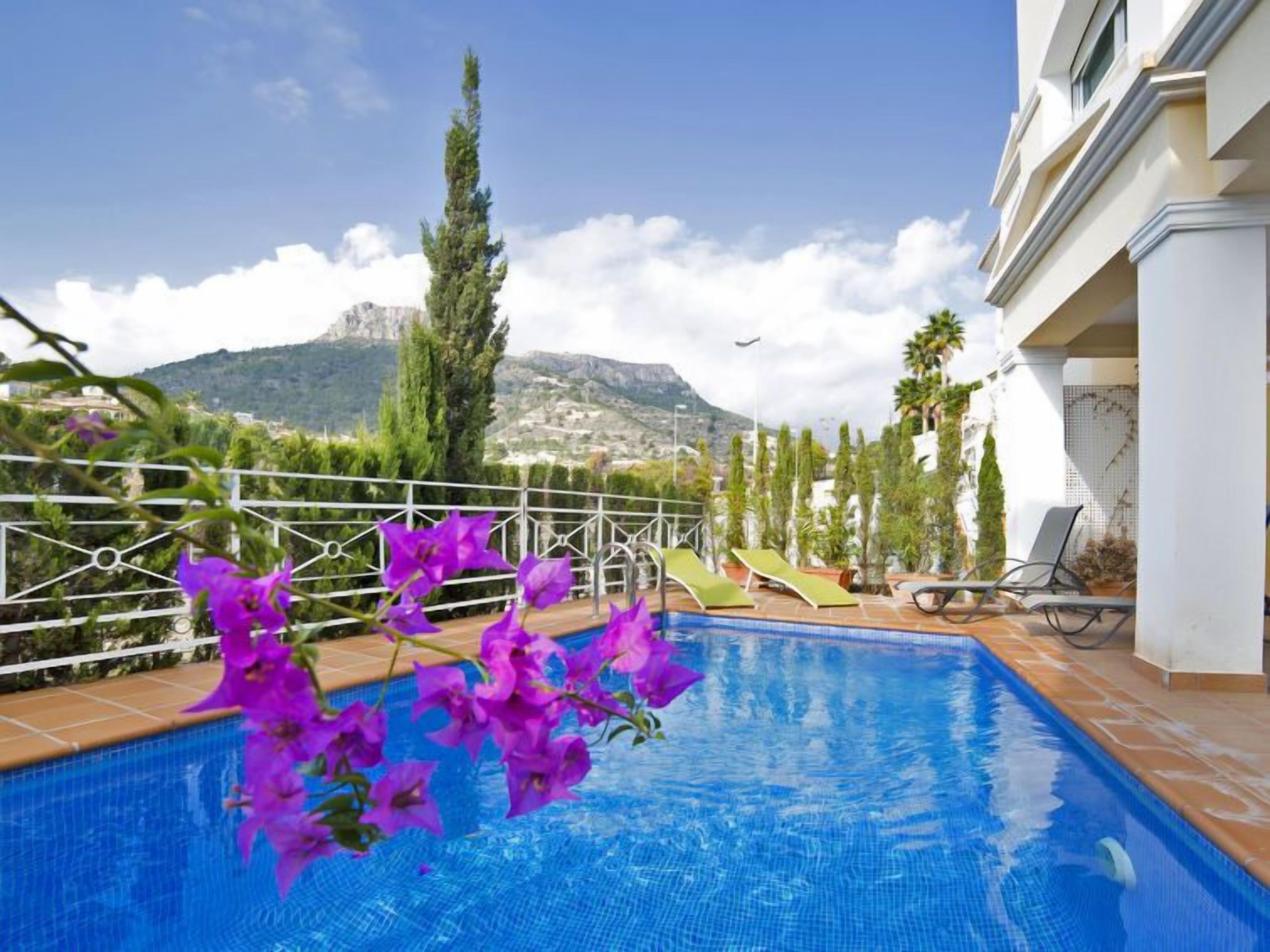 A swimming pool with a view of mountains, surrounded by greenery and a white building.