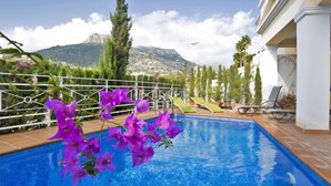 Piscina con vista sulle montagne, immersa nel verde e affiancata da un edificio bianco.