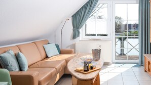 Ein modernes Wohnzimmer mit einer beigen Ledersofa, einem Holztisch und Blick durch eine Glastür auf einen Außenbereich.