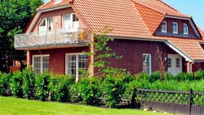 Ein zweistöckiges Backsteinhhaus mit rotem Ziegeldach, einem Balkon mit Glasgeländer und einem gepflegten Garten.