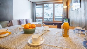 A dining area with a table set for two, a bowl of oranges, a glass of orange juice, and a window with a snowy view. - Le Grand Tichot A et B (Val Claret) by Interhome (Tignes)