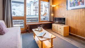 A hotel room with a bed, a wooden desk, a TV, and large windows with a snowy mountain view. - Le Grand Tichot A et B (Val Claret) by Interhome (Tignes)