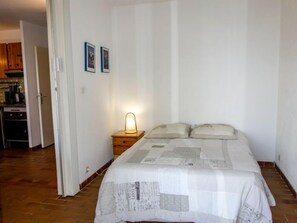 A bedroom with a bed, a nightstand with a lamp, and a kitchen visible through an open door.
