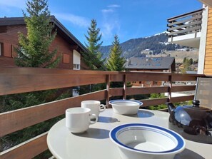 Una terraza de madera con una mesa preparada para dos, con vista a un paisaje montañoso con casas y árboles.