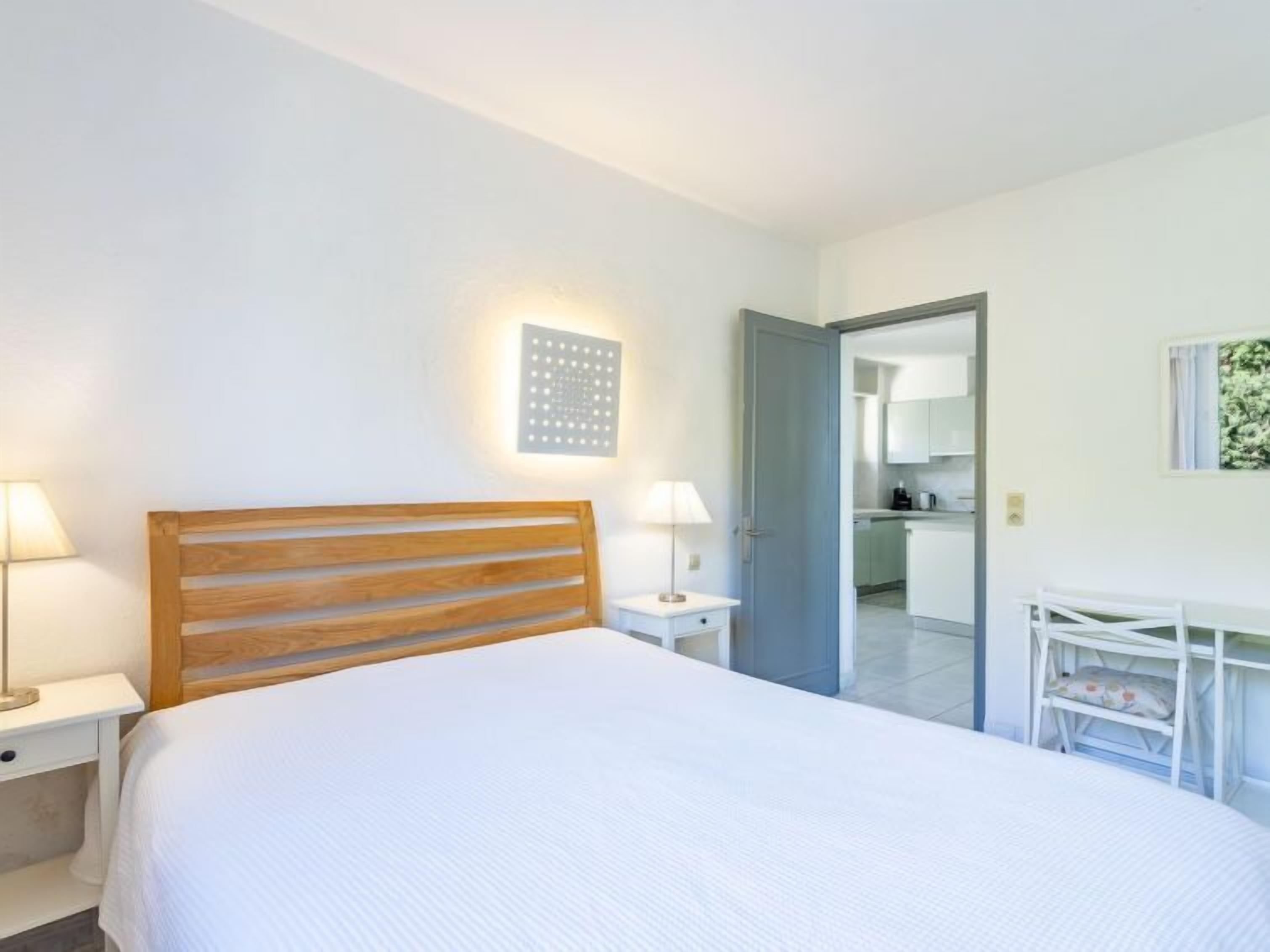 A bedroom with a wooden headboard, a bedside table with a lamp, a small desk with a chair, and a view of the kitchen through an open door.