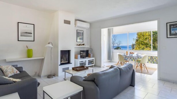 A modern living room with a grey sofa, a white coffee table, and a TV on a white stand. There is a balcony with a table and chairs, and a view of the sea. - Carpe Diem by Interhome (Rayol Canadel)