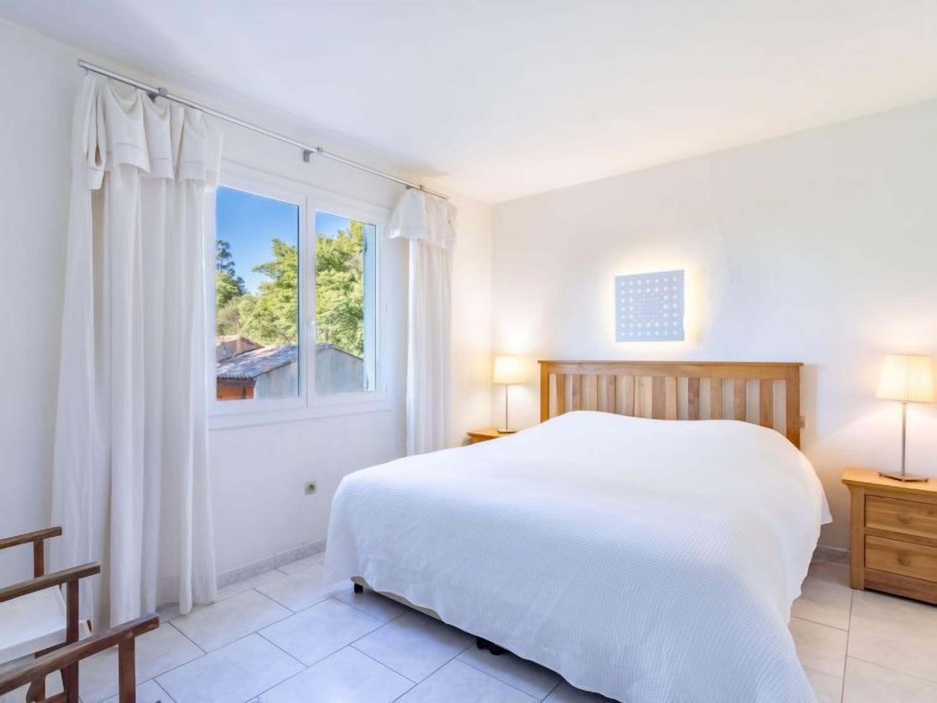A bedroom with a large bed, two bedside tables, a window with curtains, and a wall-mounted light fixture.