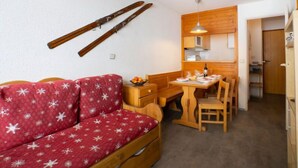 A cozy room with a red sofa, wooden dining table, and chairs. There is a kitchen area with wooden cabinets and a hanging light fixture. - Le Hameau du Borsat (Val Claret) by Interhome (Tignes)