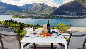 Une table avec une bouteille de vin, un verre, du pain et des tomates, placée sur un balcon donnant sur un lac et des montagnes.