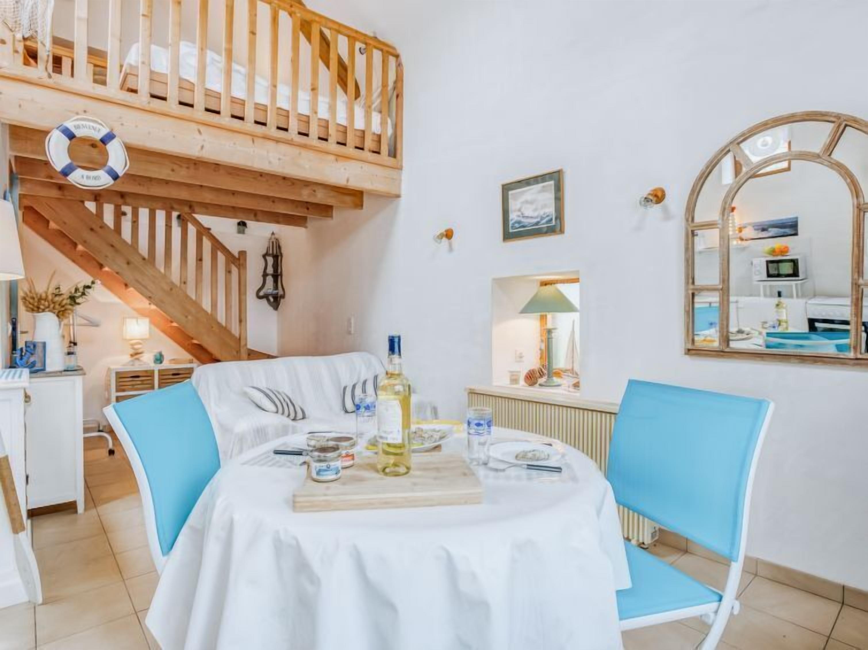 Un coin repas avec un escalier en bois, une table blanche recouverte d’une nappe blanche, des chaises bleues et une planche en bois.