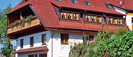 Ein zweistöckiges Haus mit rotem Dach, zahlreichen Fenstern und Balkonen mit Pflanzen.