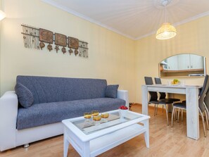 Uma sala de estar com um sofá cinza, uma mesa de centro branca com bebidas, uma área de jantar com mesa e cadeiras, e uma cozinha com armários e um espelho.