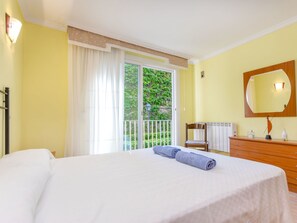 Quarto de hotel com cama, cómoda de madeira, espelho e vista para vegetação através de uma porta de vidro deslizante.