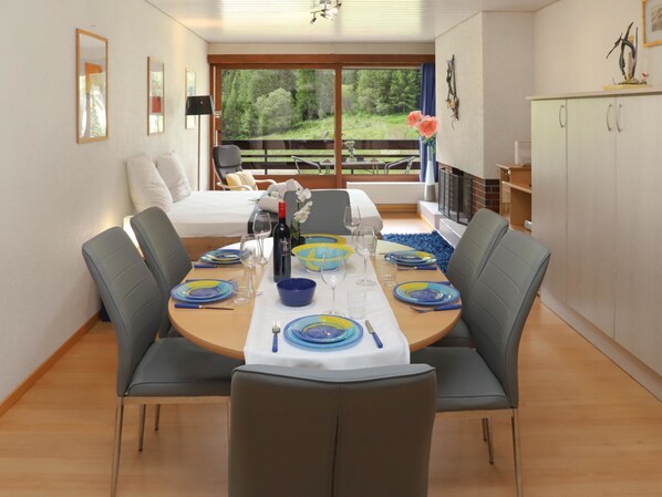 Zona de dinar amb taula rodona ben a punt per a sis persones, envoltada de cadires grises. Es veuen un llit i un sofà al fons, amb vista a arbres a través de grans finestres.