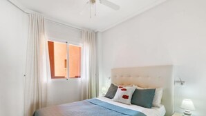 Un dormitorio con cama, cortinas, una ventana, una mesita de noche y una lámpara.