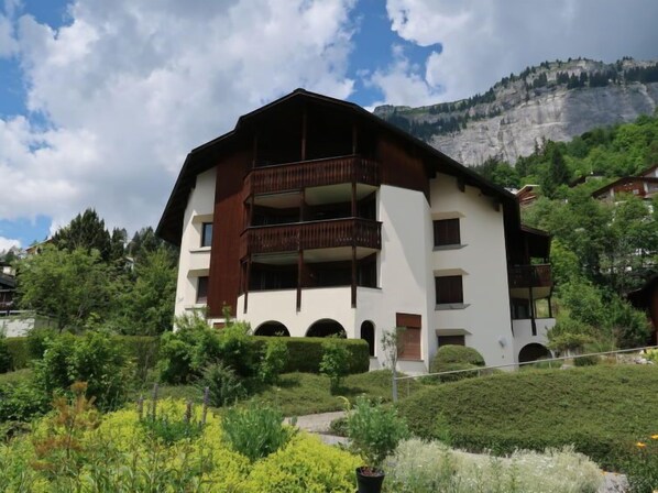A multi-story building with balconies, surrounded by lush greenery and a mountainous backdrop.