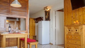 A compact kitchen with wooden cabinetry, a white refrigerator, a dining table with chairs, and a television mounted on the wall.