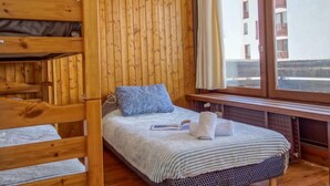 A room with a bunk bed, a single bed with a blue pillow, a window with curtains, and a view of buildings outside.