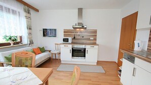 A compact kitchen with a microwave, oven, and coffee maker. There is a dining area with a table and chairs, and a small seating area with a sofa and cushions. - Christl by Interhome (Fügen)