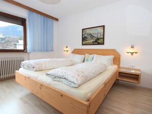 Un lit double avec une literie à motifs floraux, une tête de lit en bois, deux tables de chevet avec des lampes murales, une fenêtre donnant sur les montagnes et un radiateur.