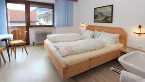 A bedroom with a wooden bed, two bedside tables, a window with curtains, a radiator, a chair, and a bathtub. - Christl by Interhome (Fügen)