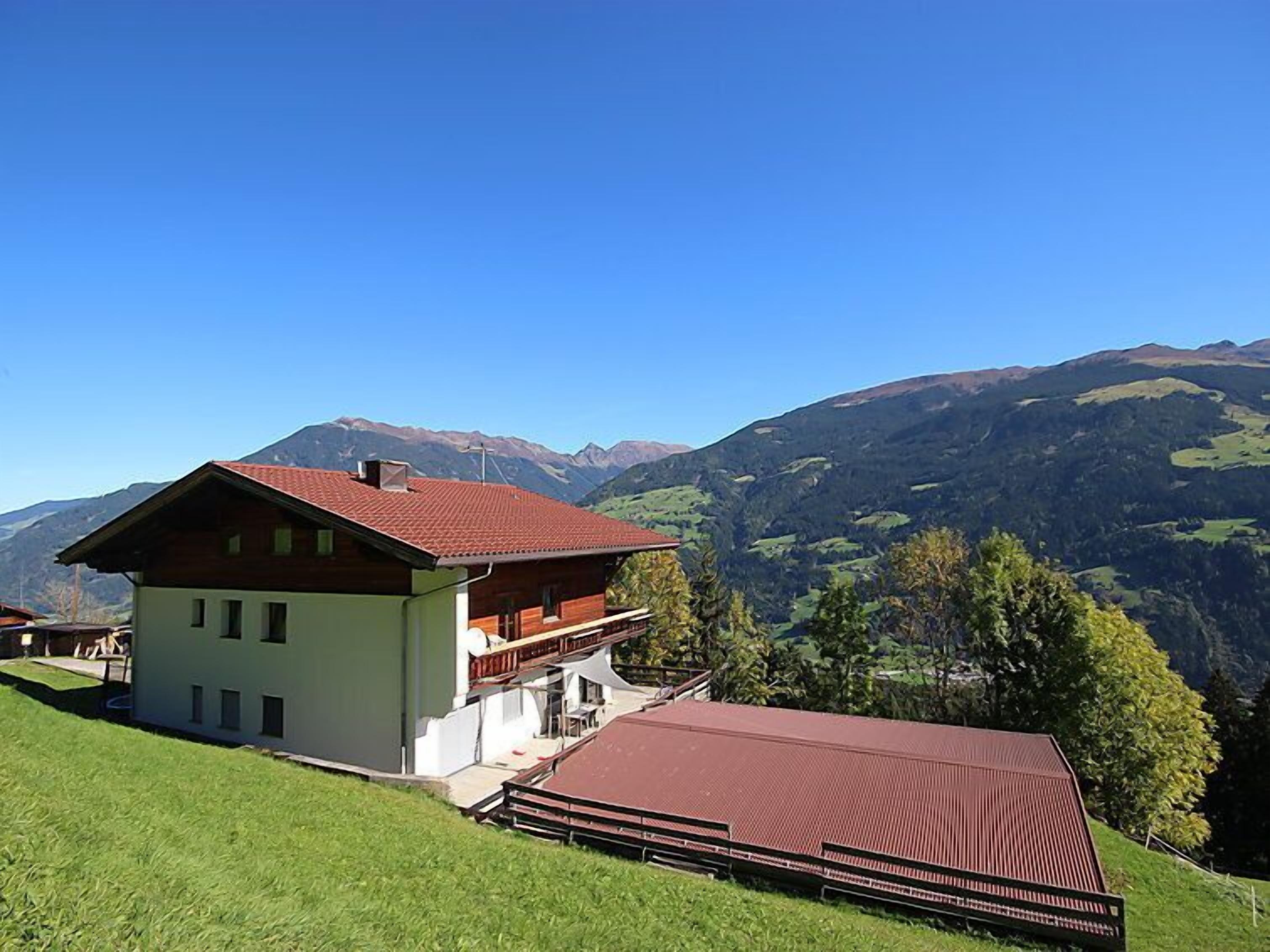 A two-story chalet with a red roof, situated on a grassy hillside with a mountain backdrop.
