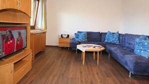 Un espacio habitable pequeño con un armario de madera, un sofá azul con cojines estampados, una mesita de madera y un televisor sobre un mueble de madera.