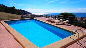 A rectangular swimming pool with a clear blue water surface, surrounded by a tiled deck. There are lounge chairs aligned along the pool's edge, and a metal railing separates the pool from the deck. The pool is set against a backdrop of a clear sky and a distant view of a coastline. - Las Alondras by Interhome (Roses)