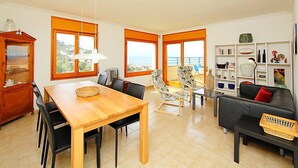 A spacious living area with a wooden dining table, multiple chairs, a black sofa, and a bookshelf. Large windows offer a view of the sea. - Las Alondras by Interhome (Roses)