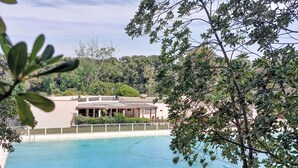 Une piscine entourée d’une clôture blanche, bordée de verdure, et un bâtiment avec un toit recouvert de tuiles.