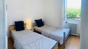 Quarto com duas camas de solteiro, janela com vista, radiador e luminária suspensa.