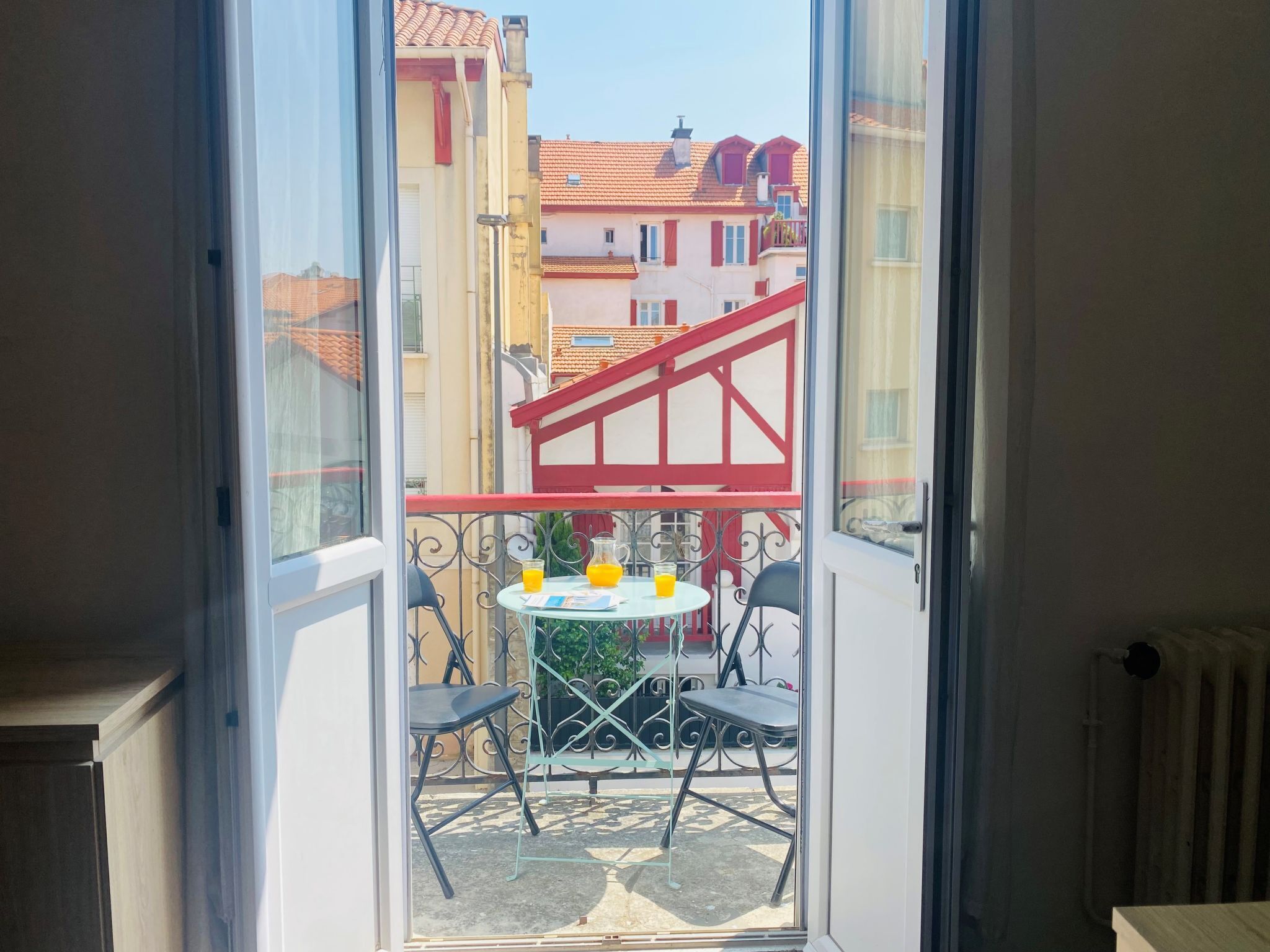Un balcon avec une table et des chaises, une rambarde rouge et une vue sur les bâtiments voisins.
