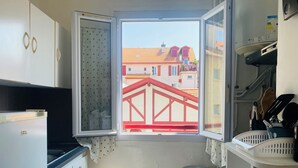 Uma cozinha compacta com vista para um edifício de telhado vermelho, uma lixeira de aço inoxidável e uma geladeira branca.