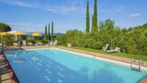 A swimming pool with lounge chairs and umbrellas, surrounded by greenery and tall trees. - Apartment Lucia by Interhome (Bucine)
