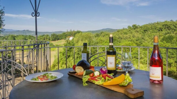 A table with a bottle of wine, a glass, and a plate of salad on a balcony with a scenic view. - Apartment Lucia by Interhome (Bucine)
