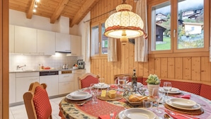 Een eetgedeelte met een ronde tafel gedekt voor zes personen, omringd door houten stoelen met rode kussens. Een kroonluchter hangt boven de tafel, en een raam biedt uitzicht op huizen op de achtergrond.