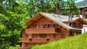 Een houten chalet met een schuin dak, meerdere balkons en een weelderige groene heuvel als achtergrond.