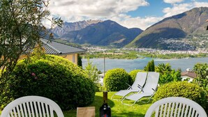 Ein Tisch mit einer Weinflasche, Gläsern und einer Tüte Snacks. Zwei weiße Stühle stehen um den Tisch herum. Im Hintergrund ist ein See und Berge zu sehen.