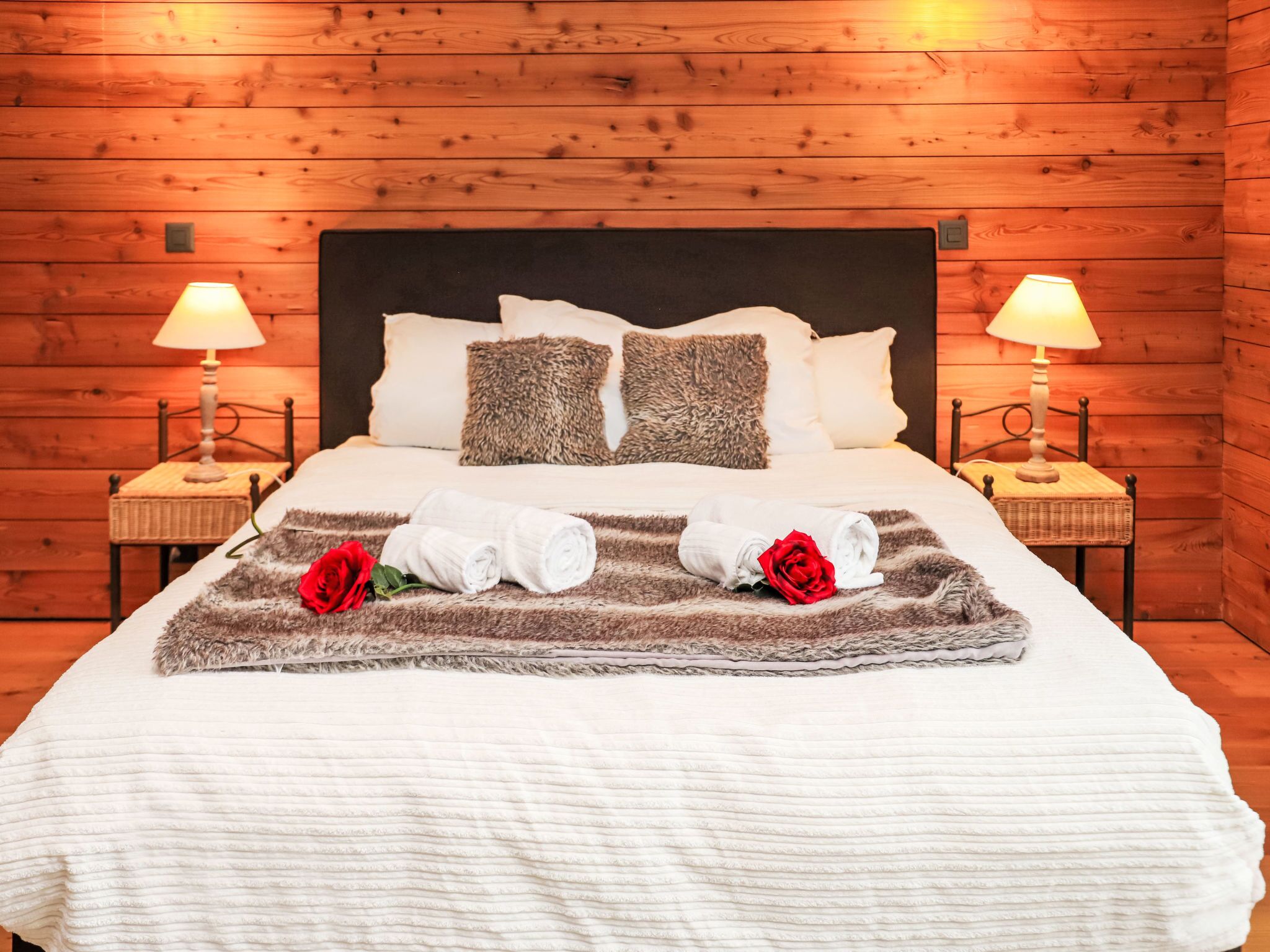 Una cama bien hecha con sábanas blancas, dos almohadas y un edredón mullido. Hay dos rosas rojas sobre el edredón. La habitación tiene paredes de madera y dos lámparas de noche.