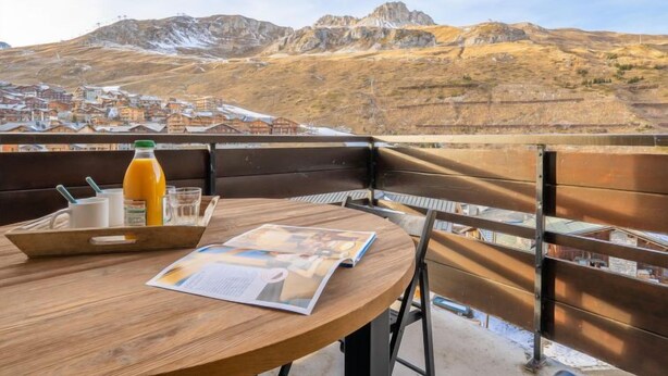 Une table en bois, sur laquelle se trouvent une bouteille de jus d’orange, des verres et un menu, est placée sur un balcon donnant sur une ville nichée dans les montagnes.