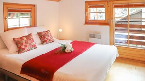 Una cama bien hecha con una colcha roja y almohadones a cuadros, una lámpara de noche y una ventana con persianas.