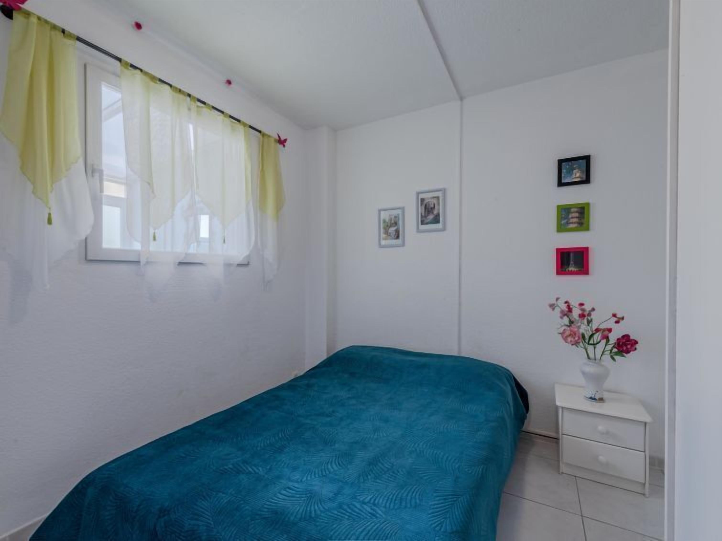 Un dormitori amb un llit individual, una tauleta de nit, una finestra amb cortines i una paret amb quadres emmarcats.