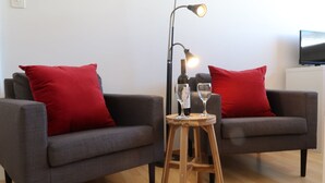 Un saló modern amb dos sofàs grisos, cadascun amb un coixí vermell, un tamboret de fusta amb dues copes de vi, i una làmpada de peu amb tres llums.