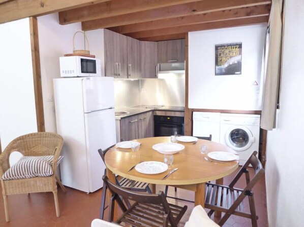 A compact kitchen with a refrigerator, microwave, stove, and washing machine. There is a dining table with plates and cutlery, and wicker chairs. - Les Cracks - Les As by Interhome (Lacanau)