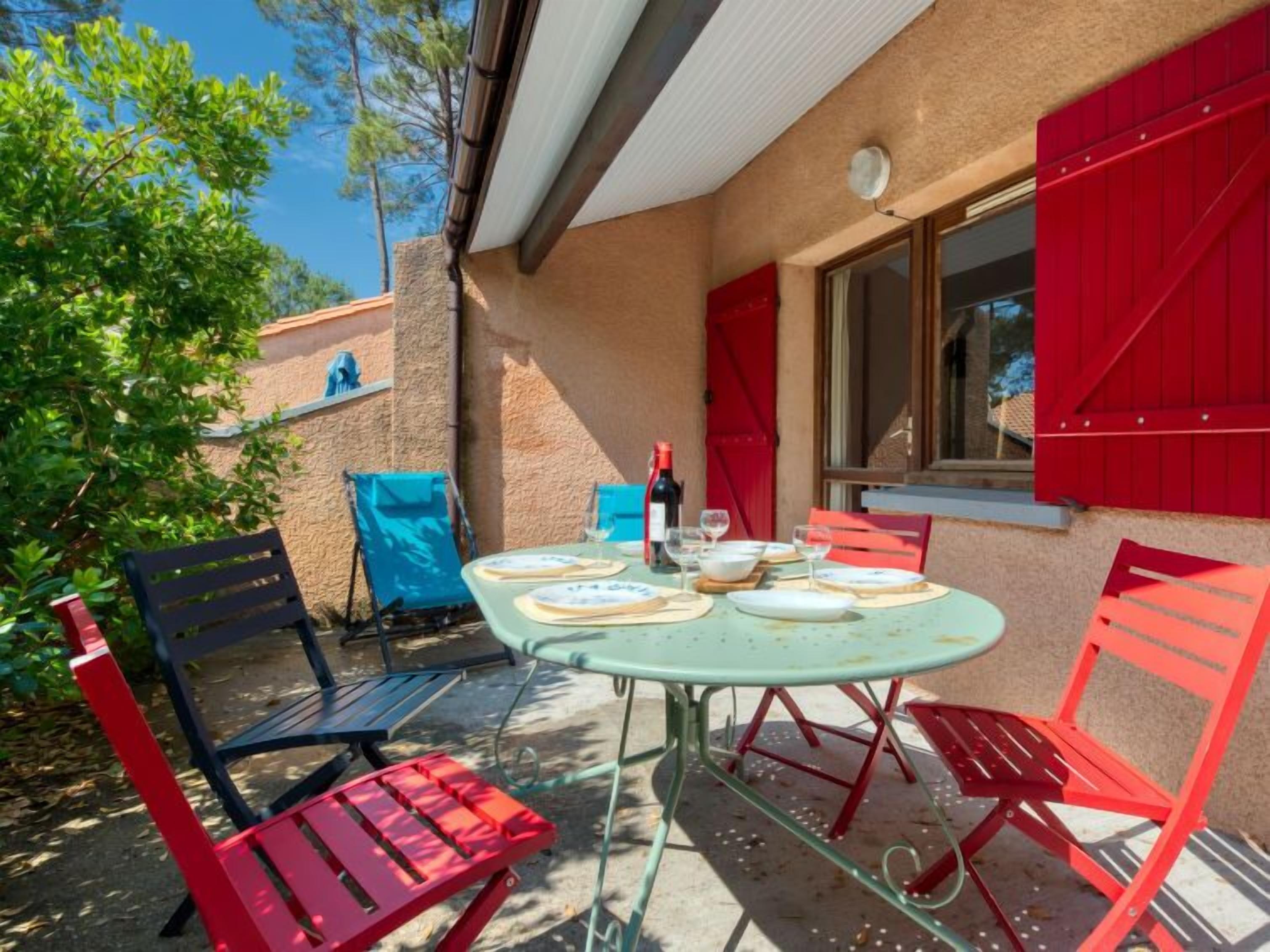 Une terrasse extérieure avec une table ronde dressée pour le repas, entourée de chaises rouges et bleues.