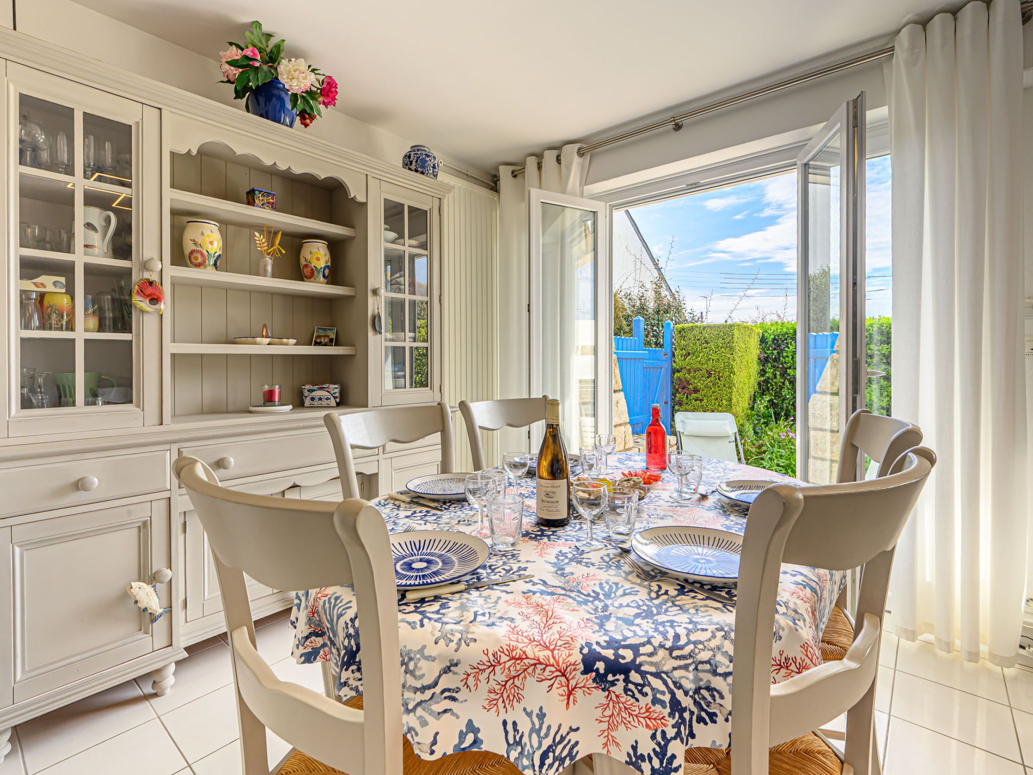 A dining area with a table set for four, surrounded by white chairs, with a view of a blue gate and greenery outside.